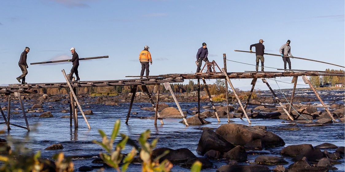 Krenkun rakentajia Tornionjoella. Valokuva, jossa kuusi henkilöä tekee puurakennelmaa kosken yllä.